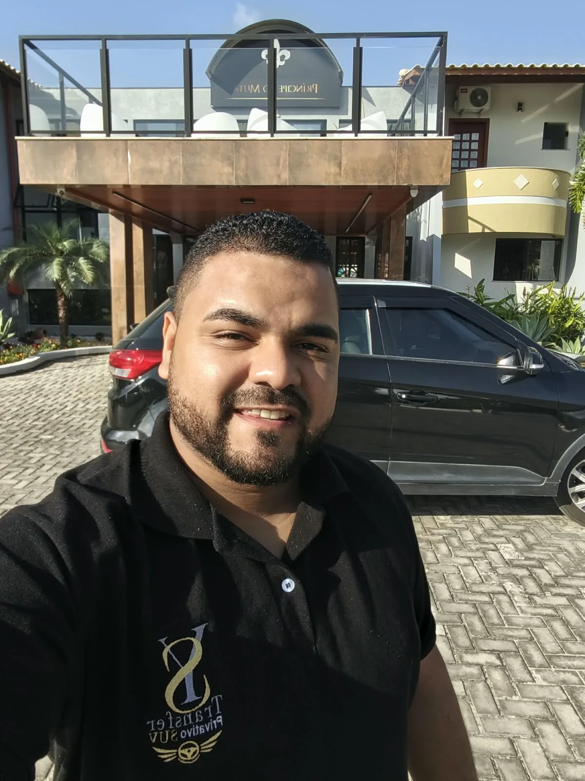 Motorista da Transfer YS uniformizado, ao lado do SUV preto em frente a uma hospedagem